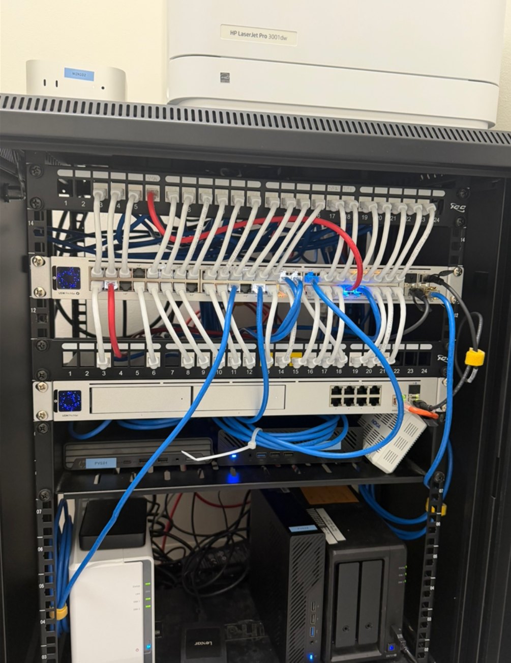 완성된 네트워크 랙. 패치 패널 두 개와 스위치에 흰색과 파란색 패치 케이블이 연결되어 있고, 아래 칸에는 미니 PC와 NAS가 있다. 랙 위에는 맥 미니와 레이저 프린터가 놓여 있다.