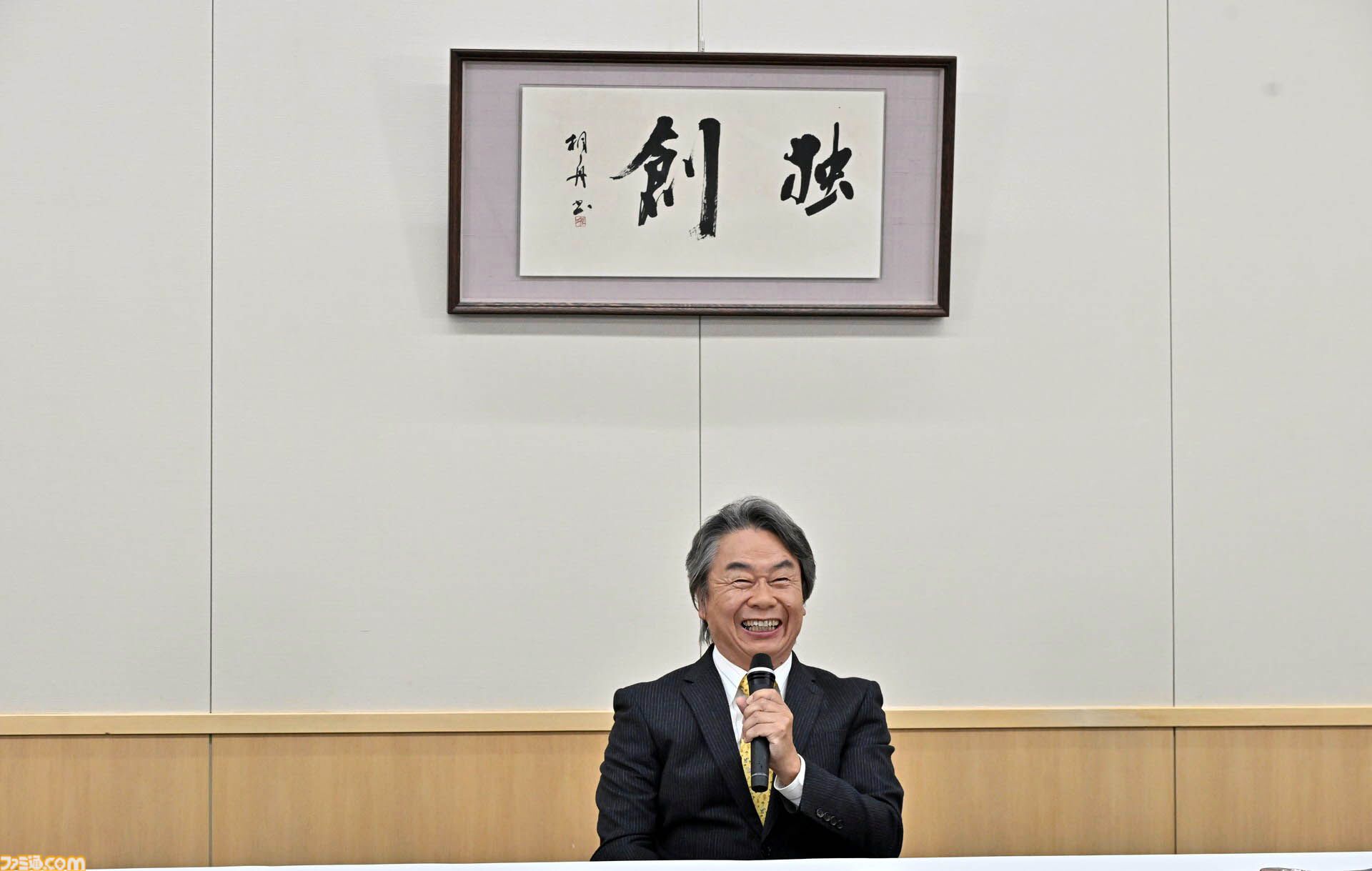 벽면의 ‘독창’ 편액 아래에 앉아 마이크를 두 손으로 쥐고 활짝 웃는 미야모토 시게루.