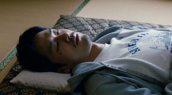 빈 방 다다미 위 이불에서 천장을 보고 누워 있는 30대 초반 일본인 남자. 회색 후드와 흰 티셔츠 차림.